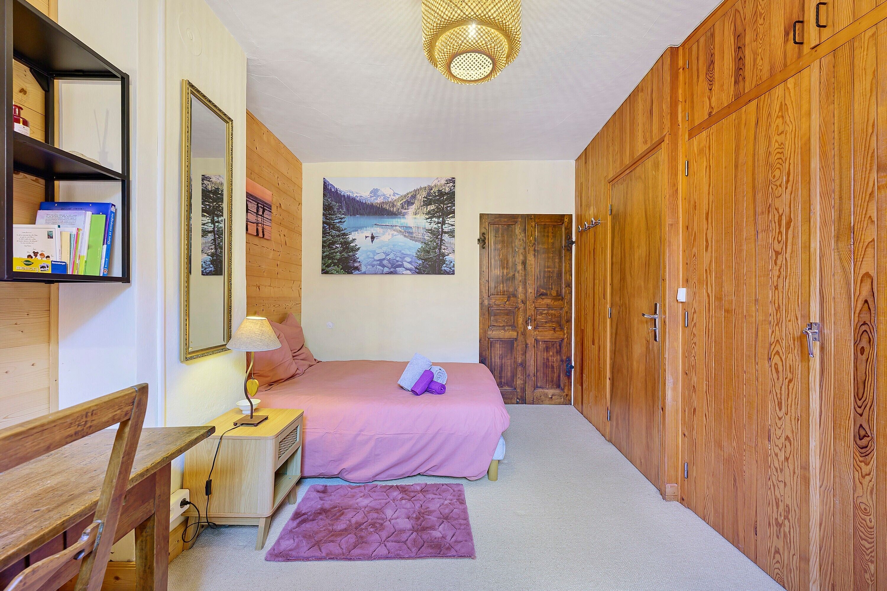 A bedroom with a bed, a desk, a chair, a mirror, a painting, and wooden wardrobes.