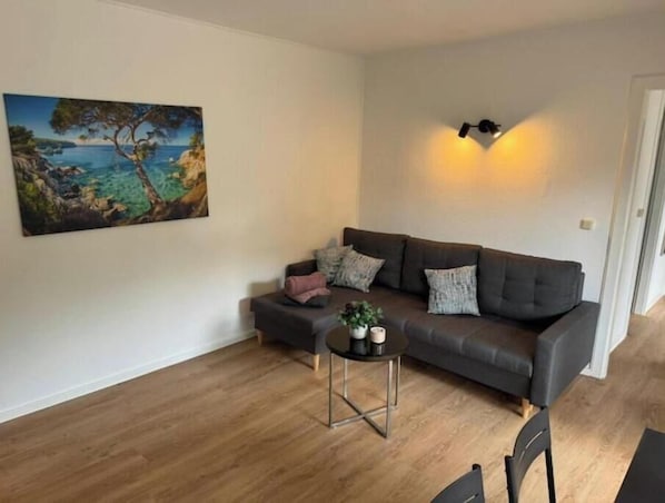 A living room with a grey sofa, a small round table with a plant, and a painting of a coastal scene on the wall.