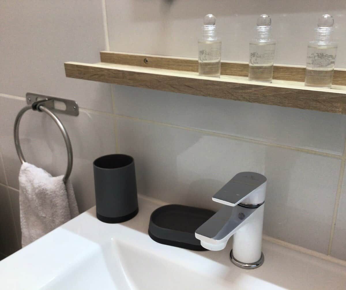 Une salle de bain avec un lavabo blanc, un porte-serviettes, une poubelle noire et une étagère sur laquelle sont posées trois bouteilles.