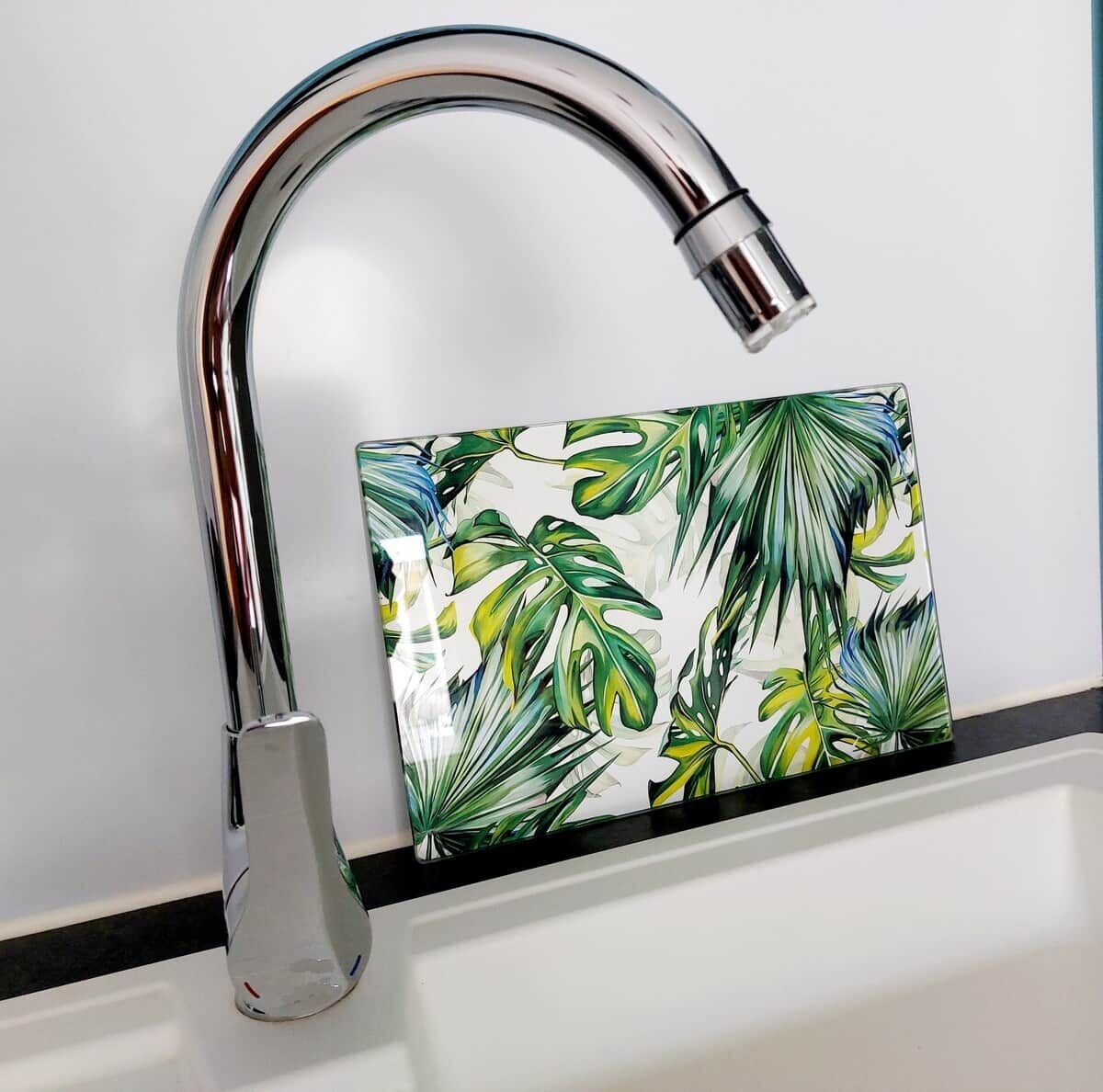 Ein chromfarbener Wasserhahn mit gebogener Ausgießer und ein weißes Waschbecken mit Rückwand in tropischem Blattmuster.