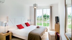 Un dormitorio con cama, lámparas de noche, televisor y ventana con vista a follaje.