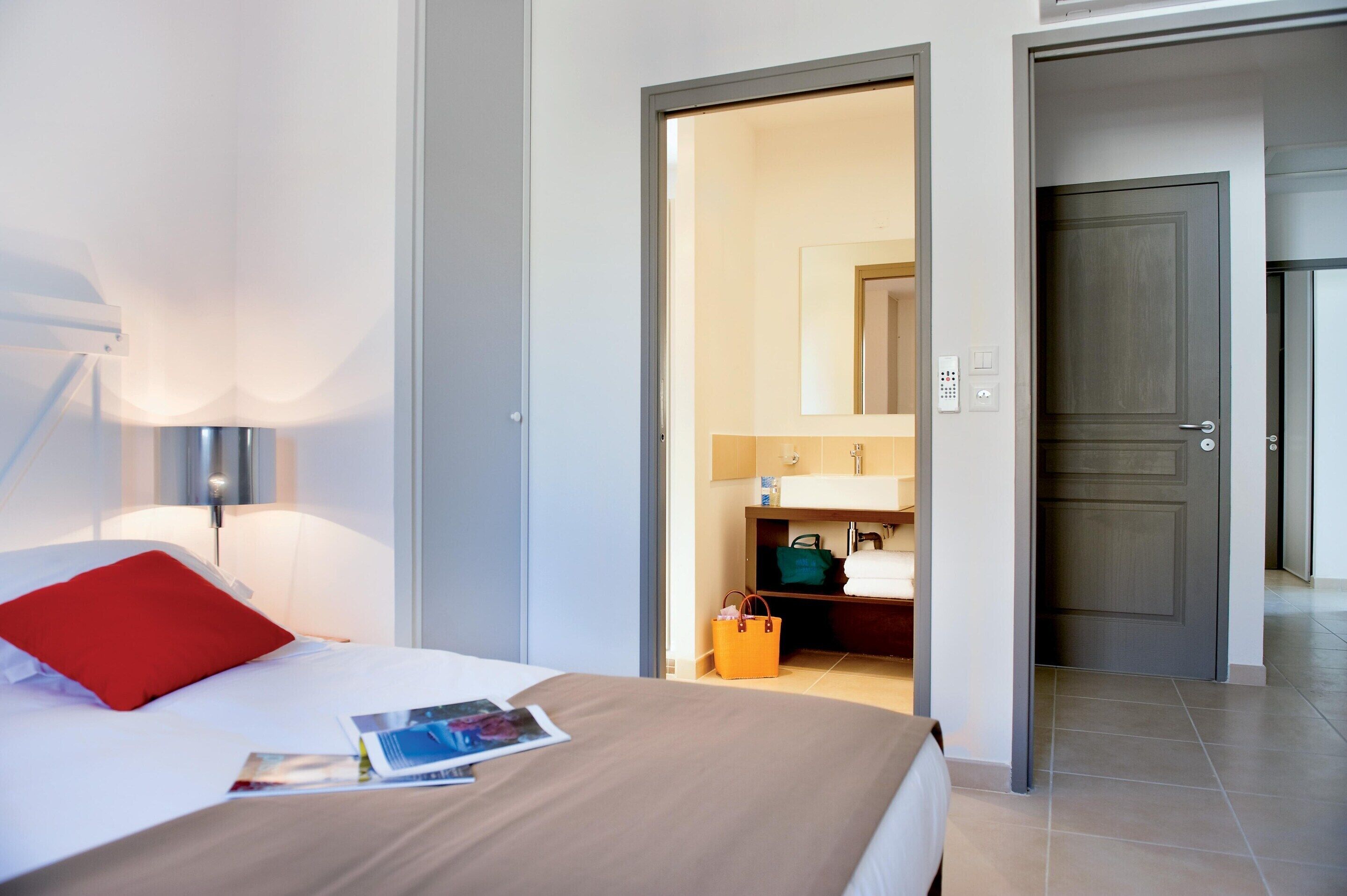 Une chambre d’hôtel comprenant un lit, une table de chevet, une lampe, une salle de bain avec un miroir et une porte donnant sur une autre pièce.