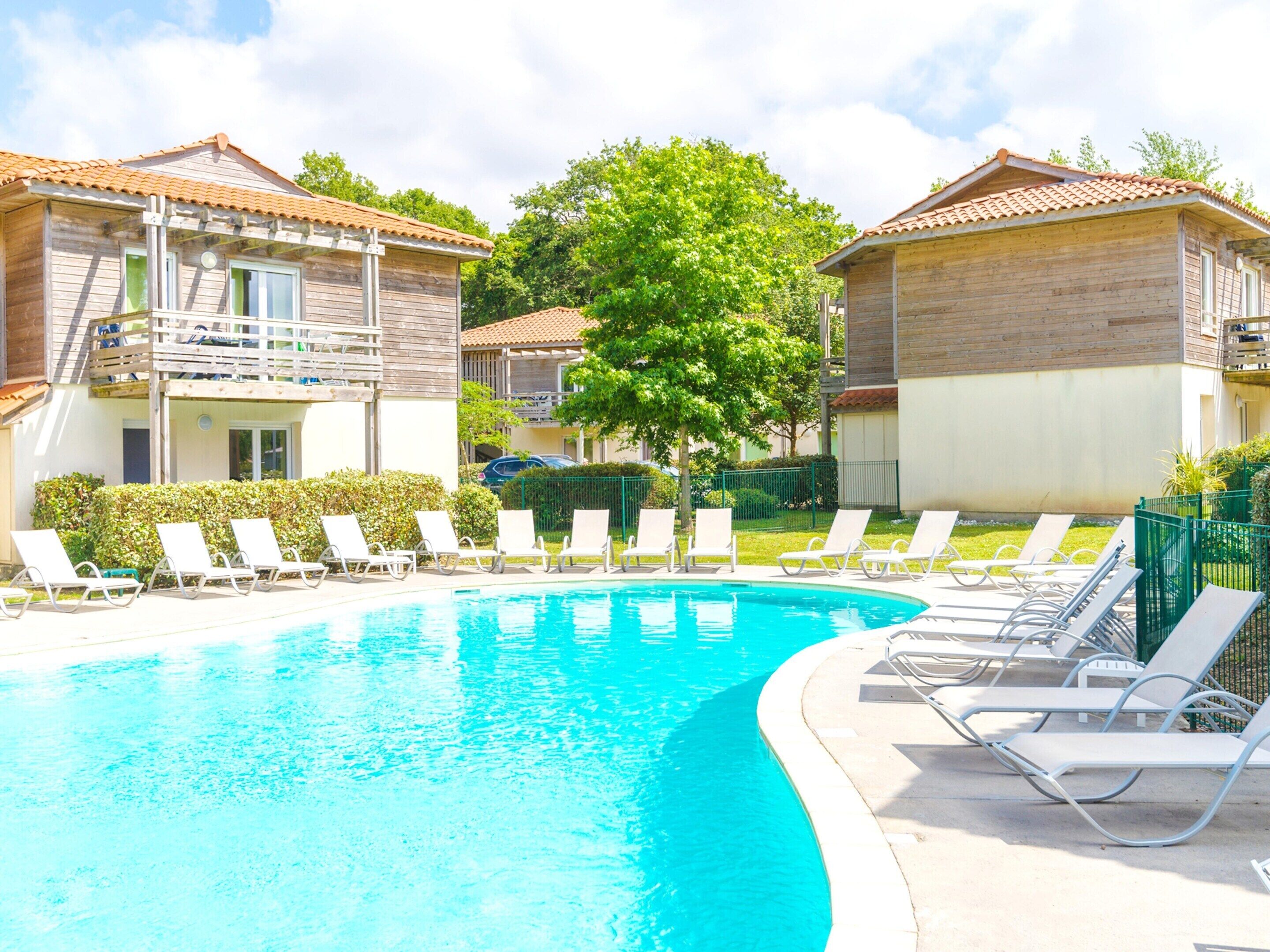 A swimming pool with lounge chairs, wooden buildings with balconies, and a tree in the center.