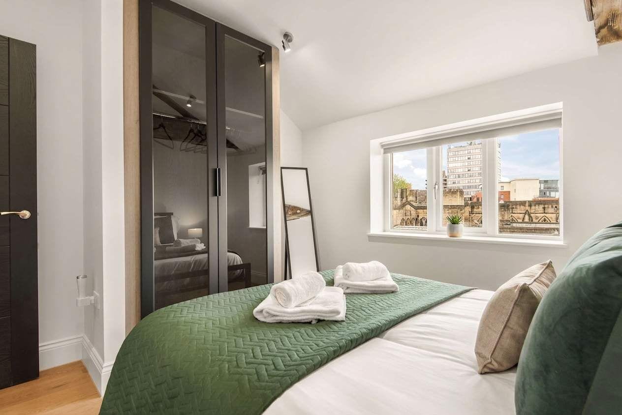 Un dormitorio con cama, ventana con vista a edificios, armario con espejo y colcha verde.
