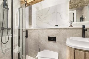 Un baño moderno con ducha de vidrio, pared con azulejos de mármol, mueble de madera y lavamanos blanco.