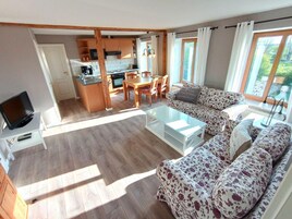 Un amplio salón con sofá, mesa de centro y televisor. Se observa una cocina con armarios de madera y un comedor con sillas.