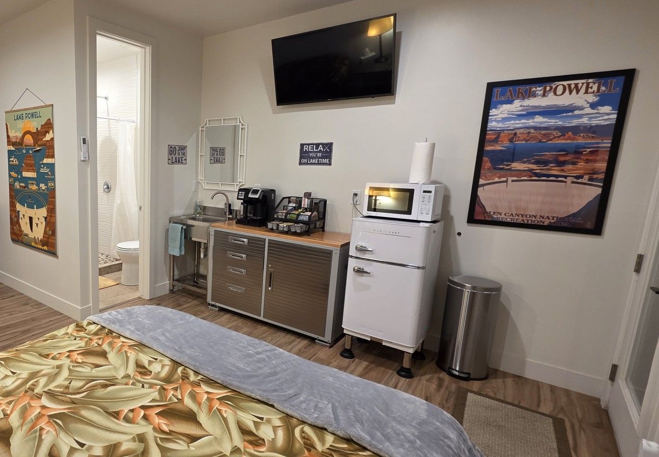 Una camera d'albergo con un letto, angolo cottura, televisione e un poster del Lago Powell, oltre a un ingresso che conduce al bagno.