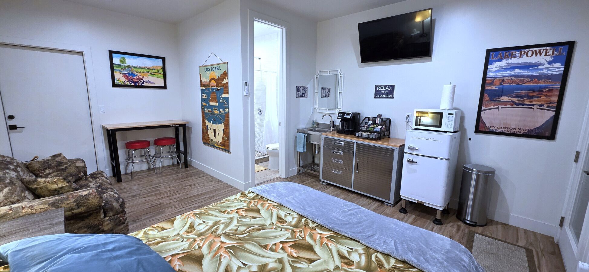 A room with a bed, a kitchenette, a TV, and a poster of Lake Powell.