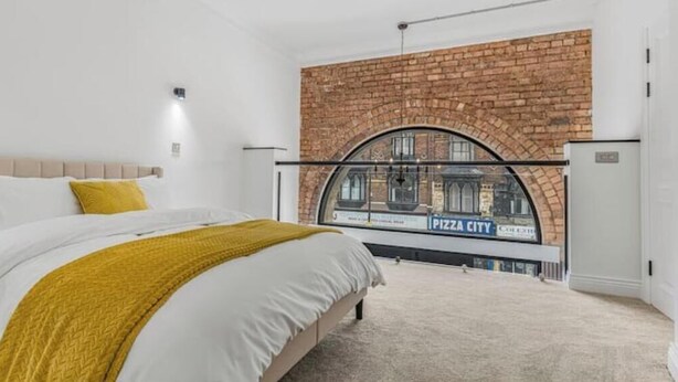 Une chambre avec un lit, un plaid jaune, un mur en briques et une fenêtre donnant sur un immeuble portant l’inscription « PIZZA CITY ».