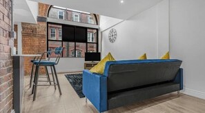 A modern living room with a blue sofa, yellow cushions, a black TV stand, and a brick wall. - Spacious 2-BedRoom Luxury Apartment in Jewellery Quarter – Central Birmingham (Birmingham)