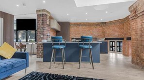 Ein modernes Wohnzimmer mit einem blauen Sofa, einem Couchtisch und einem gemusterten Teppich. Es gibt eine Küchenzeile mit einer Ziegelwand, einer Bar und zwei blauen Barhocker.