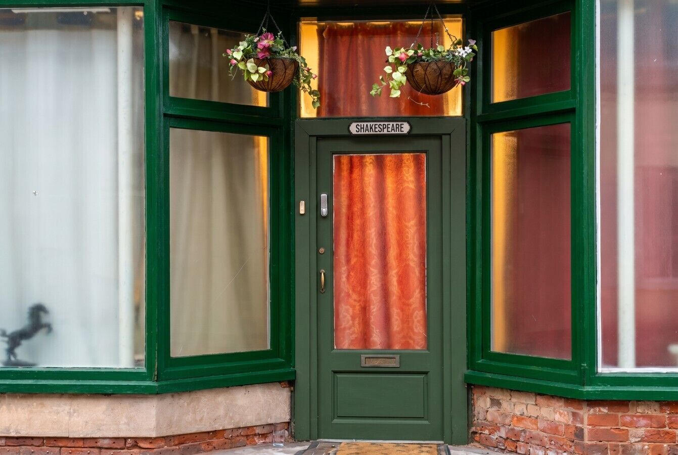 A green door with a 'Shakespeare' sign, a brick base, and two hanging flower baskets.