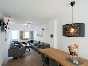 A modern living room with a wooden dining table, grey sofas, a stone accent wall, and a hanging pendant light.