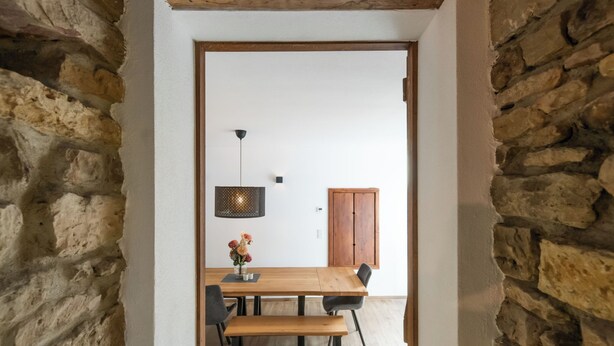 Porta in pietra che conduce a una sala da pranzo con tavolo, sedie, una panca, un lampadario e una porta in legno.