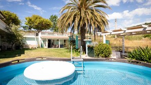 Een zwembad met een jacuzzi, omringd door een terras met een pergola, palmbomen en een huis op de achtergrond.