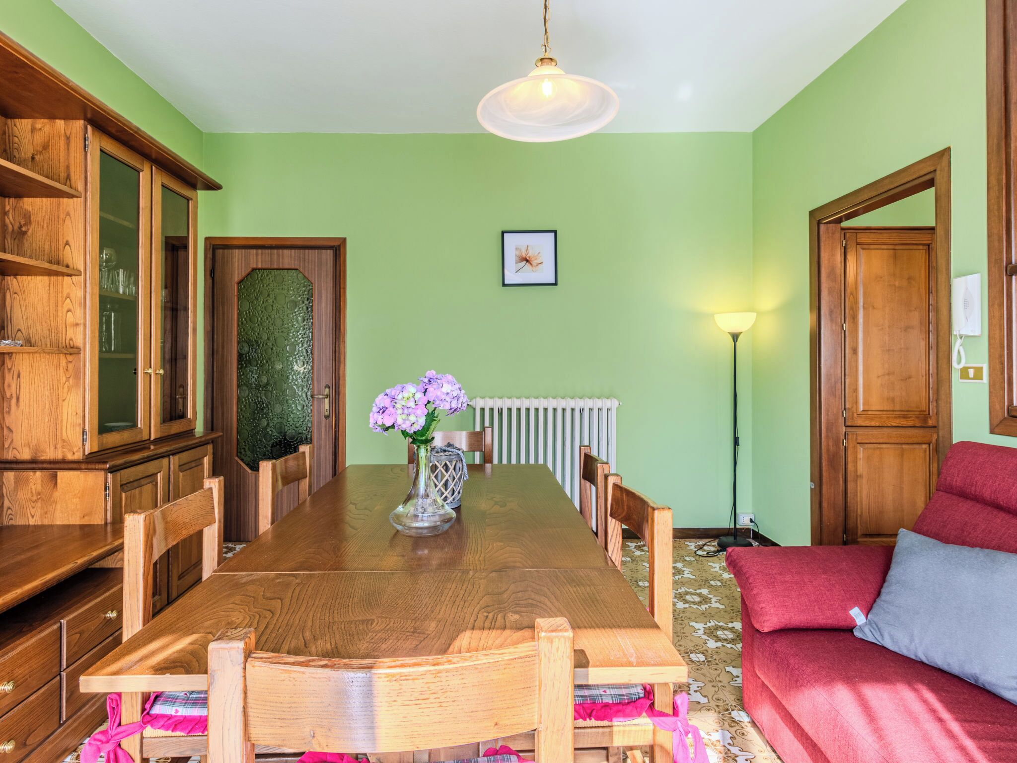 Una sala da pranzo con un tavolo di legno, sedie, un vaso con fiori, un termosifone e un divano rosso.