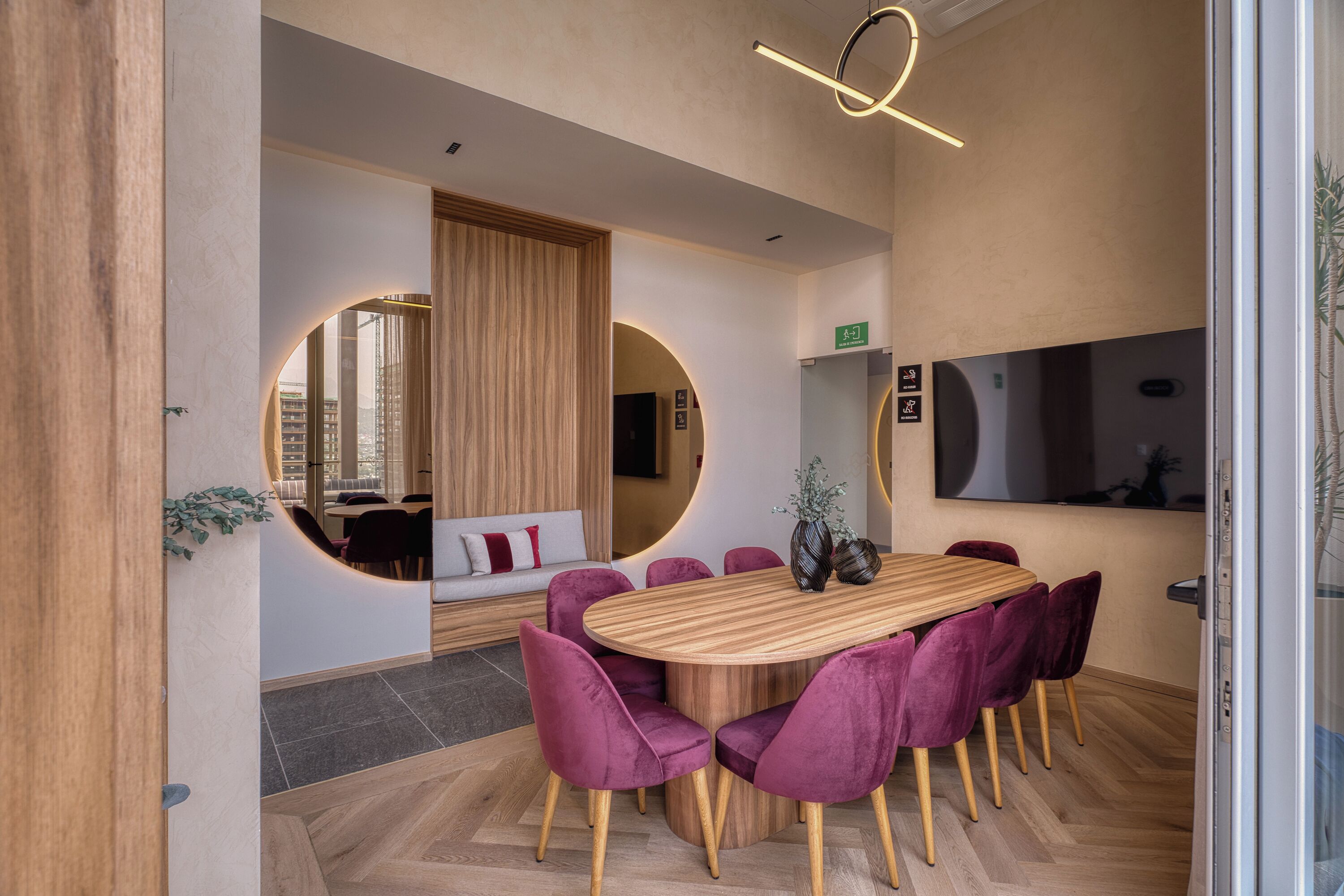Un salón de conferencias con mesa de madera, sillas moradas, un espejo grande y una lámpara moderna.