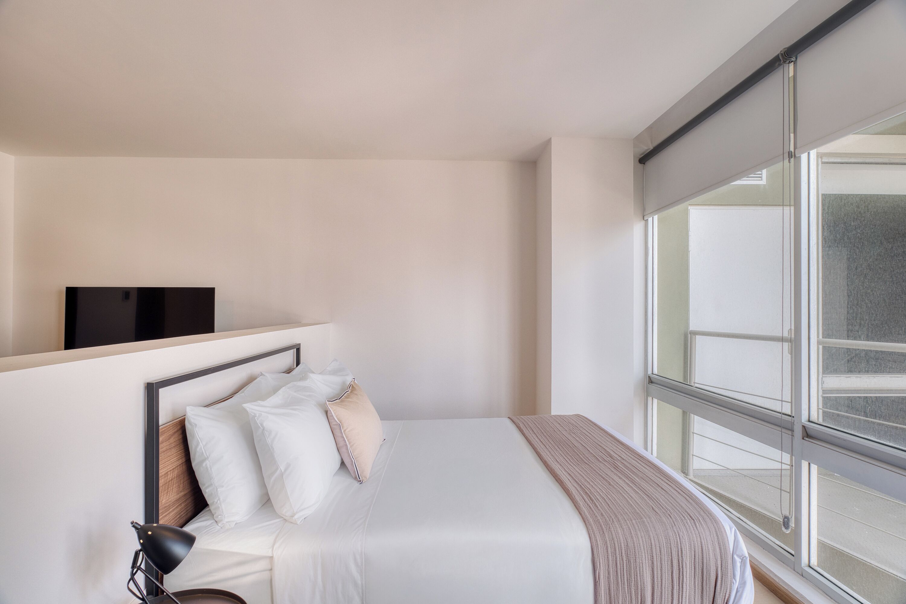 Una habitación de hotel con un ventanal grande, una cama con ropa de cama blanca, un cabecero y una mesita de noche con una lámpara.