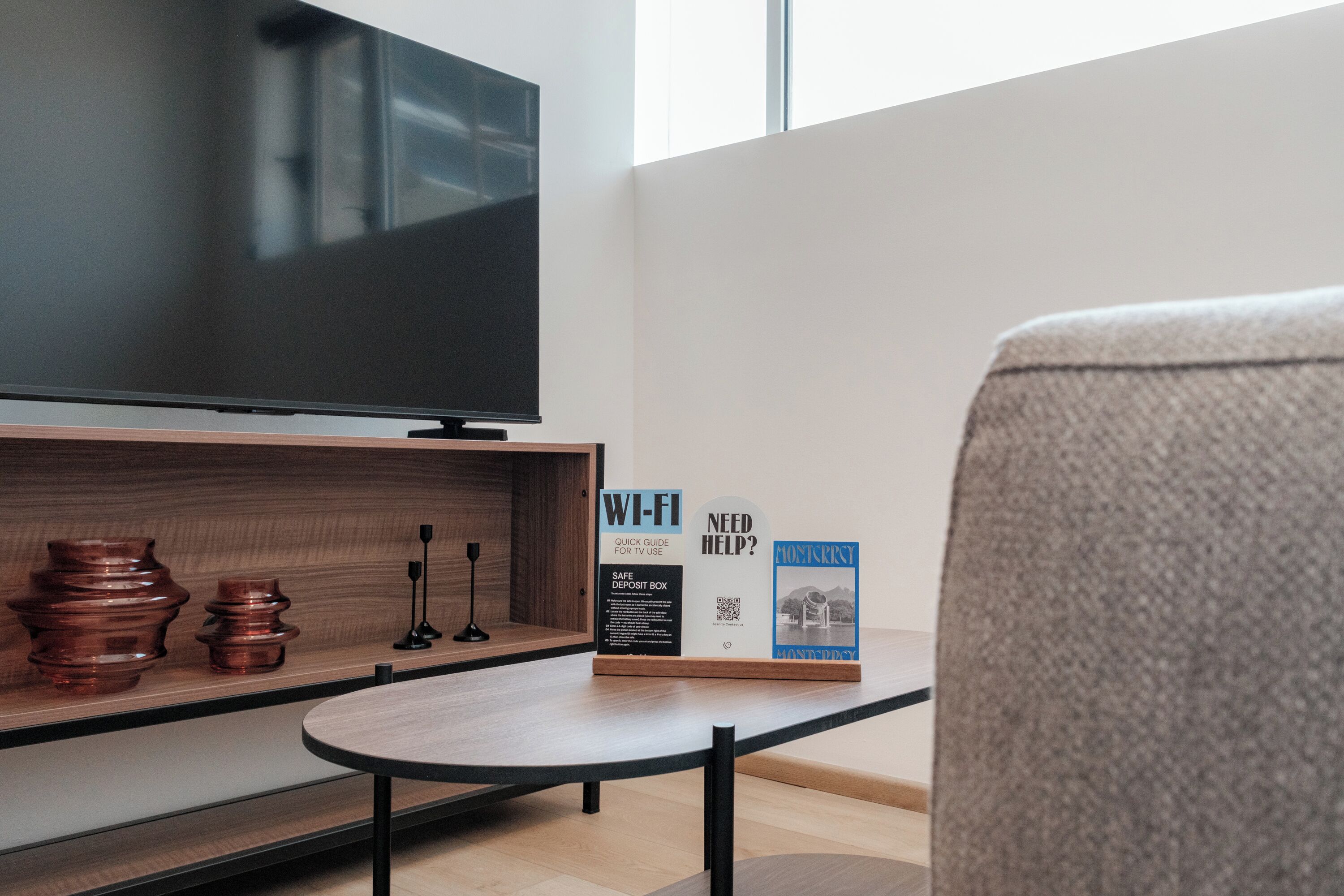Ein modernes Wohnzimmer mit Flachbildfernseher, Couchtisch und Sofa. Auf dem Couchtisch stehen dekorative Gegenstände, und es gibt ein Schild mit Informationen zu WLAN und einem Safe.