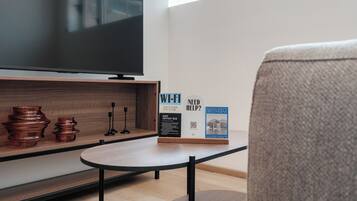 Ein modernes Wohnzimmer mit Flachbildfernseher, Couchtisch und Sofa. Auf dem Couchtisch stehen dekorative Gegenstände, und es gibt ein Schild mit Informationen zu WLAN und einem Safe.