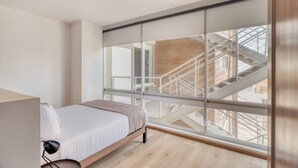 Ein Schlafzimmer mit Bett, Schreibtisch, Lampe und einem großen Fenster mit Blick auf eine Treppe.