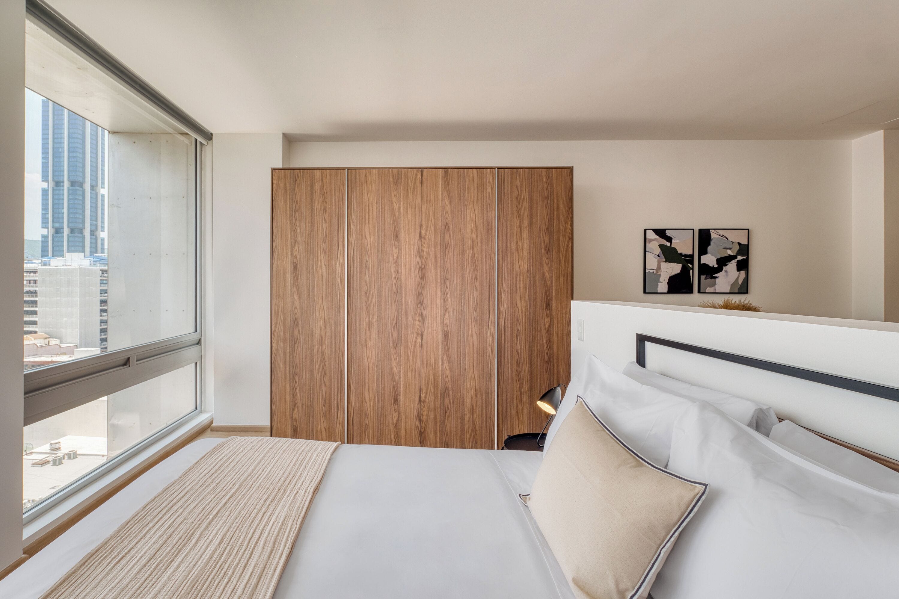 A modern hotel room with a large window, a bed with white linens, a wooden wardrobe, and abstract wall art.