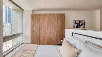 A modern hotel room with a large window, a bed with white linens, a wooden wardrobe, and abstract wall art.