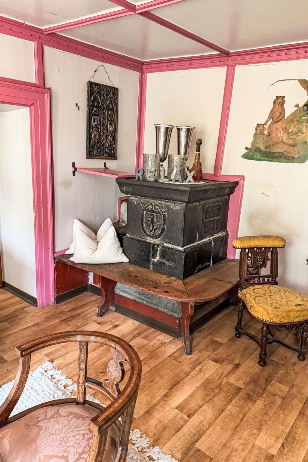 Una habitación con una estufa antigua, una silla de madera, una mesita con cojín y un cuadro en la pared.