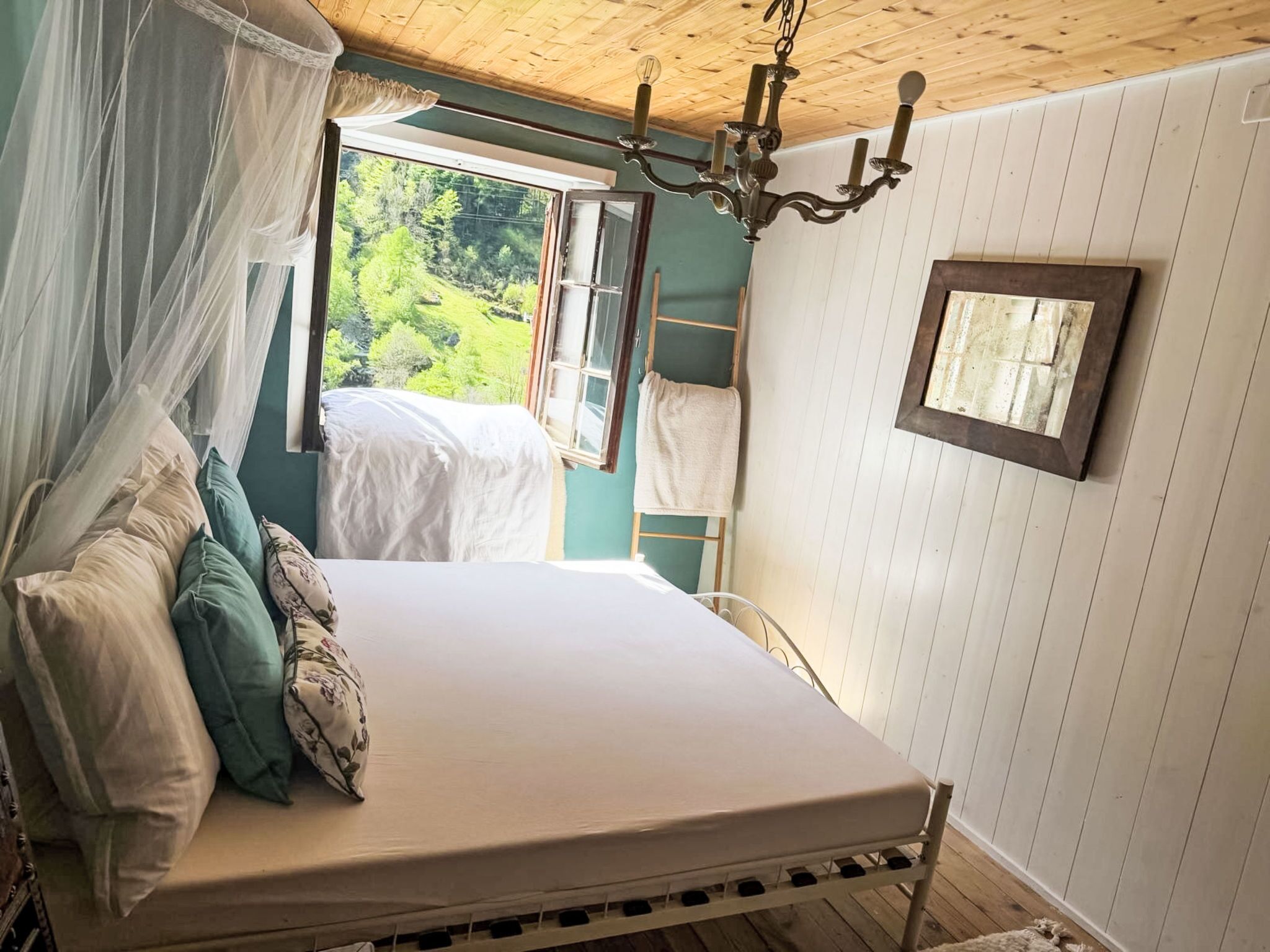 A bed with a canopy, a wooden ceiling, a chandelier, a window with a view of greenery, and a ladder against the wall.