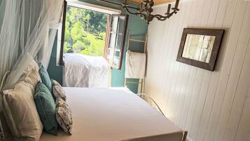 A bed with a canopy, a wooden ceiling, a chandelier, a window with a view of greenery, and a ladder against the wall.