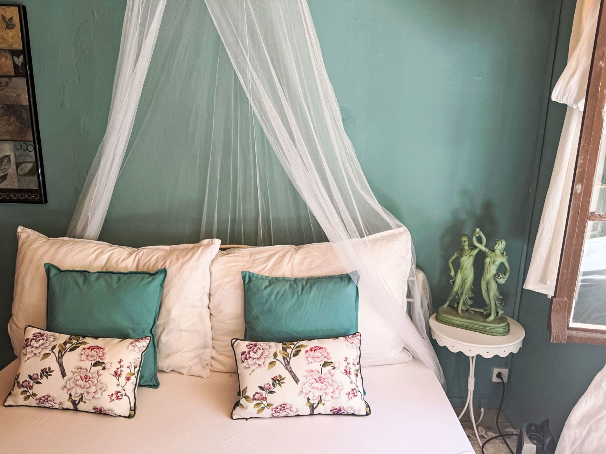 Una cama con dosel, dos almohadas, una mesita con una estatua y una pared con un cuadro enmarcado.