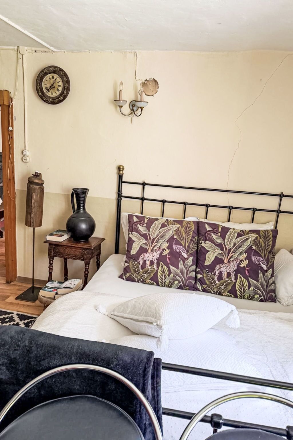 A bedroom with a metal bed frame, a wall clock, a bedside table with a vase, and a patterned pillow.