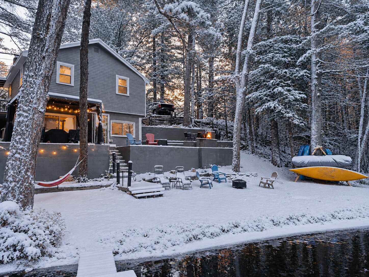 Una casa con veranda, posti a sedere all'aperto e un kayak vicino a uno stagno coperto di neve.