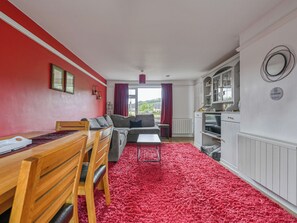 Una habitación con una mesa de comedor, sillas, un sofá, una alfombra roja, una zona de cocina y vistas a través de una ventana.