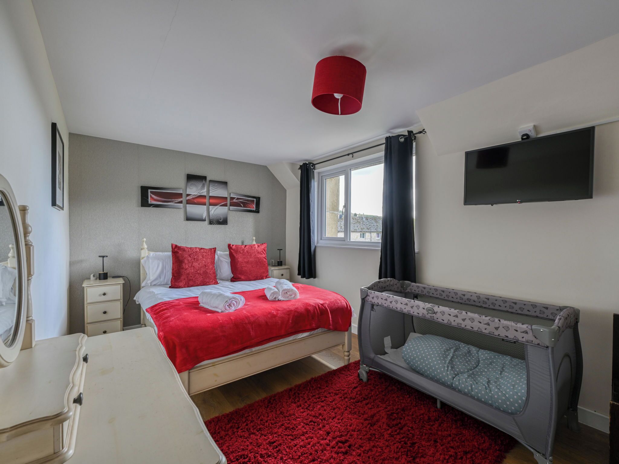 A bedroom with a bed, a dresser, a red rug, a crib, a TV, and artwork on the wall.