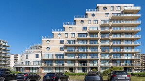 En flereetagers boligbygning med altaner, parkeringsplads og en klar blå himmel.