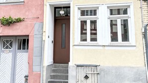 Un edifici de diverses plantes amb una porta marró i finestres d'estructura blanca, un carrer empedrat i una reixa metàl·lica.