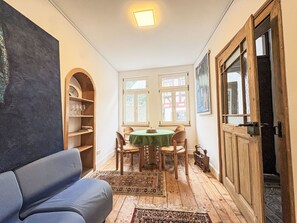 A cozy room with a wooden floor, a green table, and chairs. There's a bookshelf, a sofa, and a door with a mirror.
