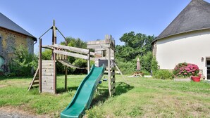 A wooden play structure with a slide in a grassy area, surrounded by buildings.