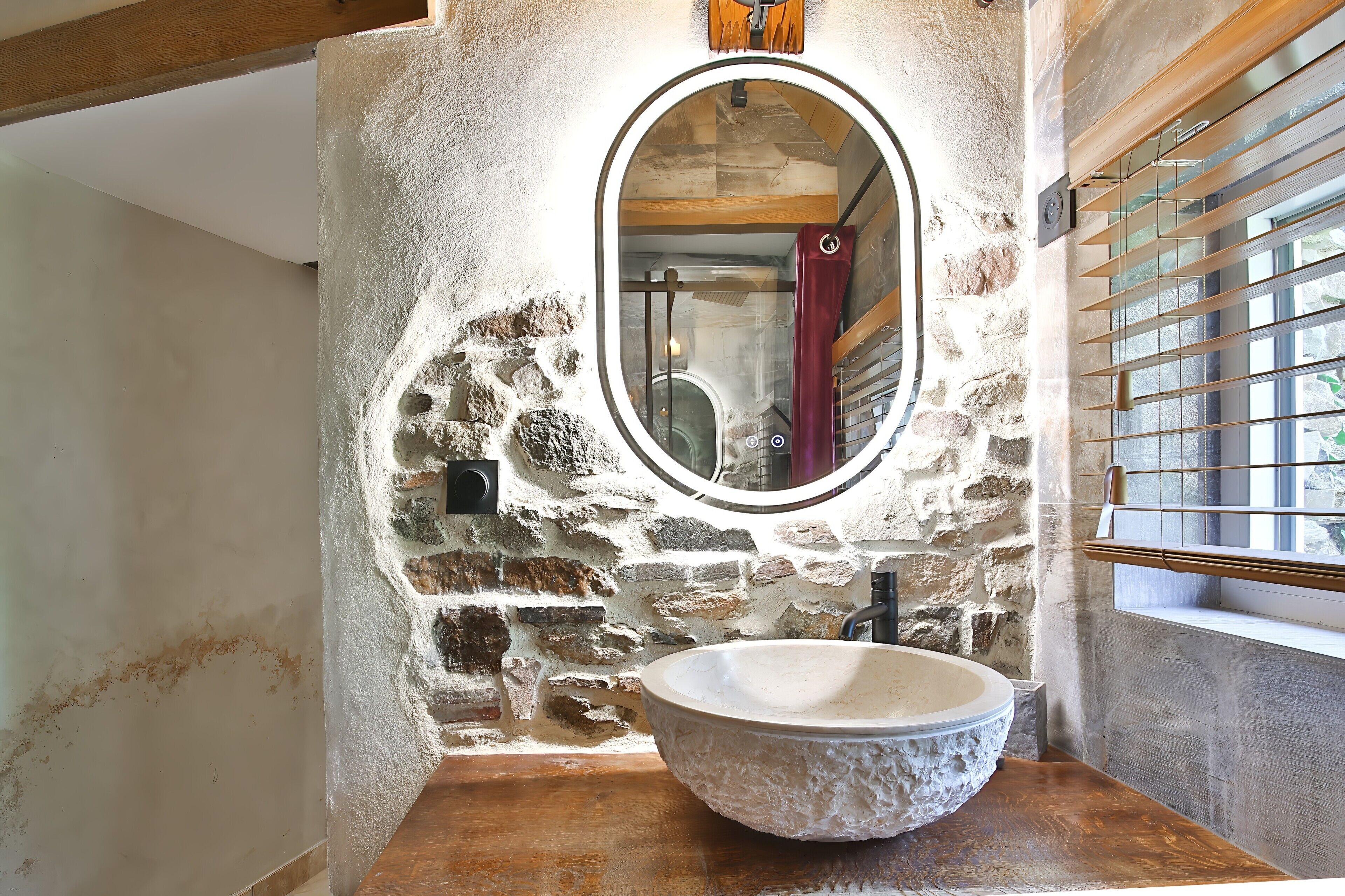 A bathroom with a stone wall, a round mirror, a wooden vanity, and a window with blinds.