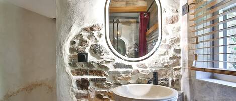 A bathroom with a stone wall, a round mirror, a wooden vanity, and a window with blinds.