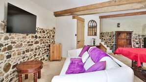A living room with a stone wall, a white sofa with purple cushions, a wooden table, and a flat-screen TV mounted on the wall.