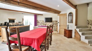 A dining area with a red tablecloth, wooden chairs, a stone wall, a television mounted on the wall, and a staircase leading to an upper level.