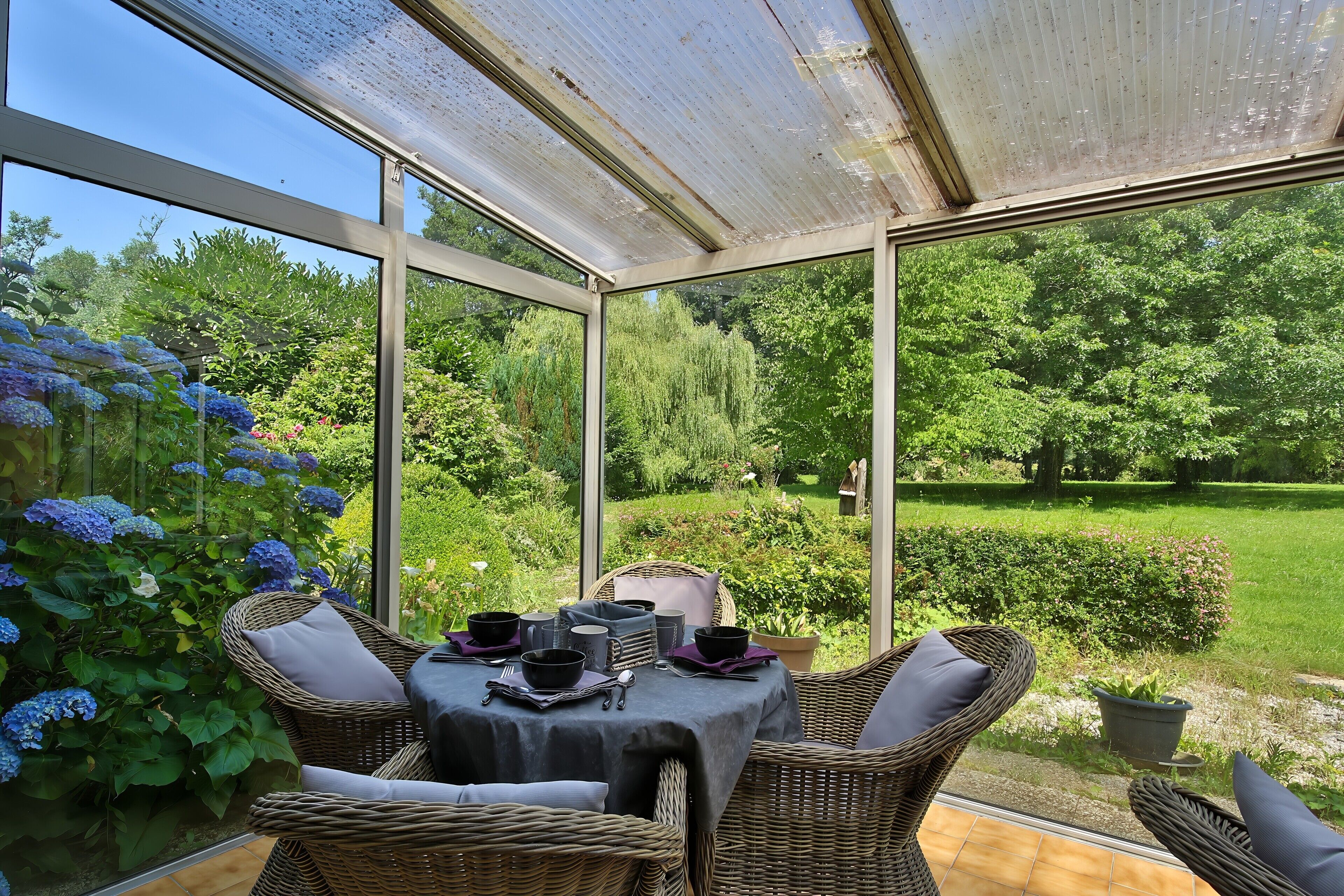 Een oranjerie met een eettafel, rieten meubels en uitzicht op een weelderige tuin.