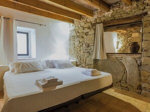 A bedroom with a stone wall, a bed with white linens, a window with curtains, and a stone fireplace. - Mas Silvestre by Interhome (La Bisbal D'Emporda)