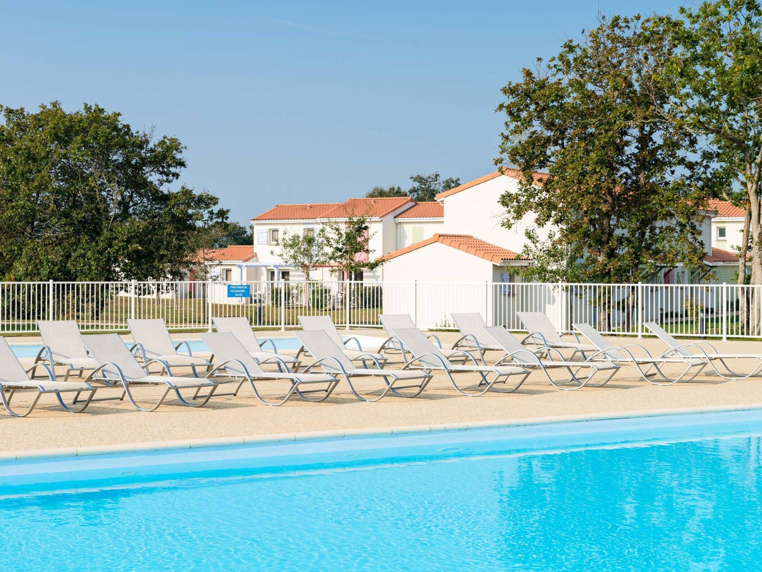 Une piscine entourée de chaises longues, une clôture blanche et, au fond, des bâtiments aux toits rouges.