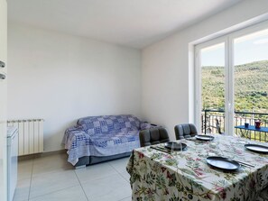 Ein Zimmer mit Esstisch für zwei Personen, einer Couch mit gemustertem Überwurf und Blick auf eine hügelige Landschaft durch ein großes Fenster.