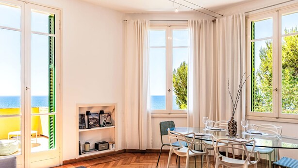 Een lichte kamer met houten vloer, een eettafel met stoelen, een bank en uitzicht op zee door grote ramen.