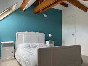 Ein Schlafzimmer mit einem weißen Bett, einem Nachttisch, einer Lampe, einer Tür und einer schrägen Decke mit Holzbalken.