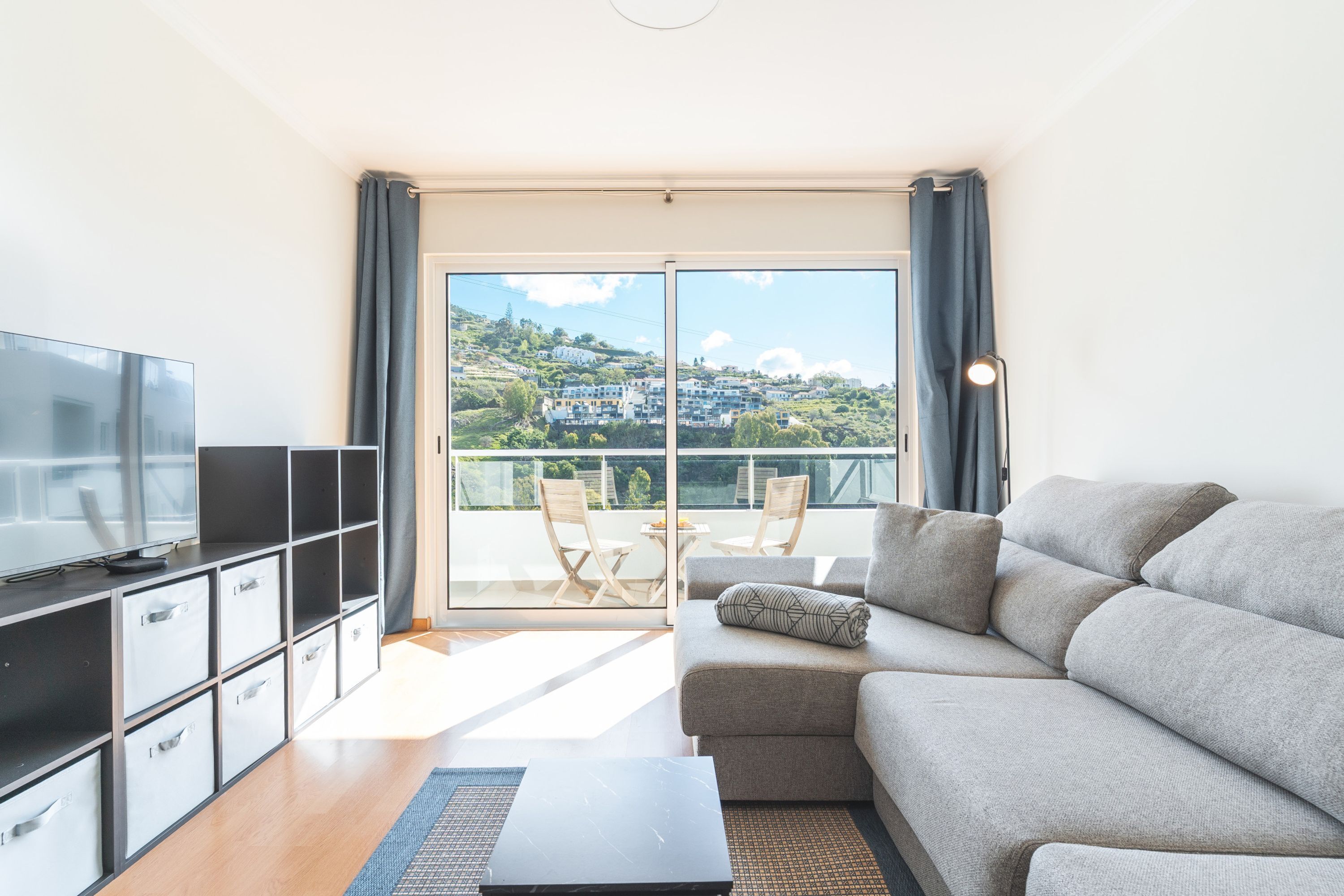 Ein modernes Wohnzimmer mit einer Couch, einem Fernseher, Stauraumeinheiten und einem Balkon mit Blick auf Gebäude.